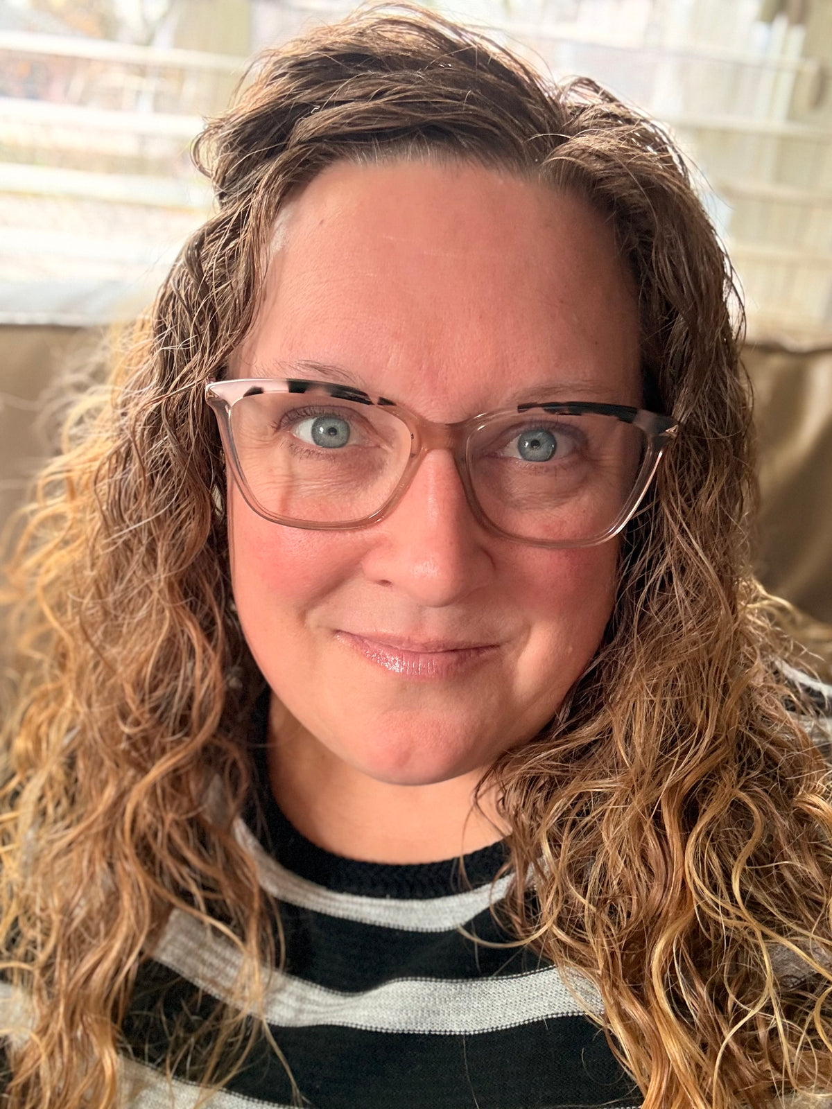 A close-up portrait of a woman with curly light brown hair wearing tortoiseshell eyeglasses and a black and white striped shirt.