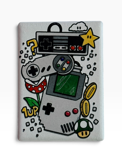 A rectangular ceramic magnet featuring a hand-painted retro video game theme. The design includes a handheld game counsel, a black controller, gray controller, gold coins, a green mushroom, a red mushroom, a yellow star, and a question mark on a light gray background.