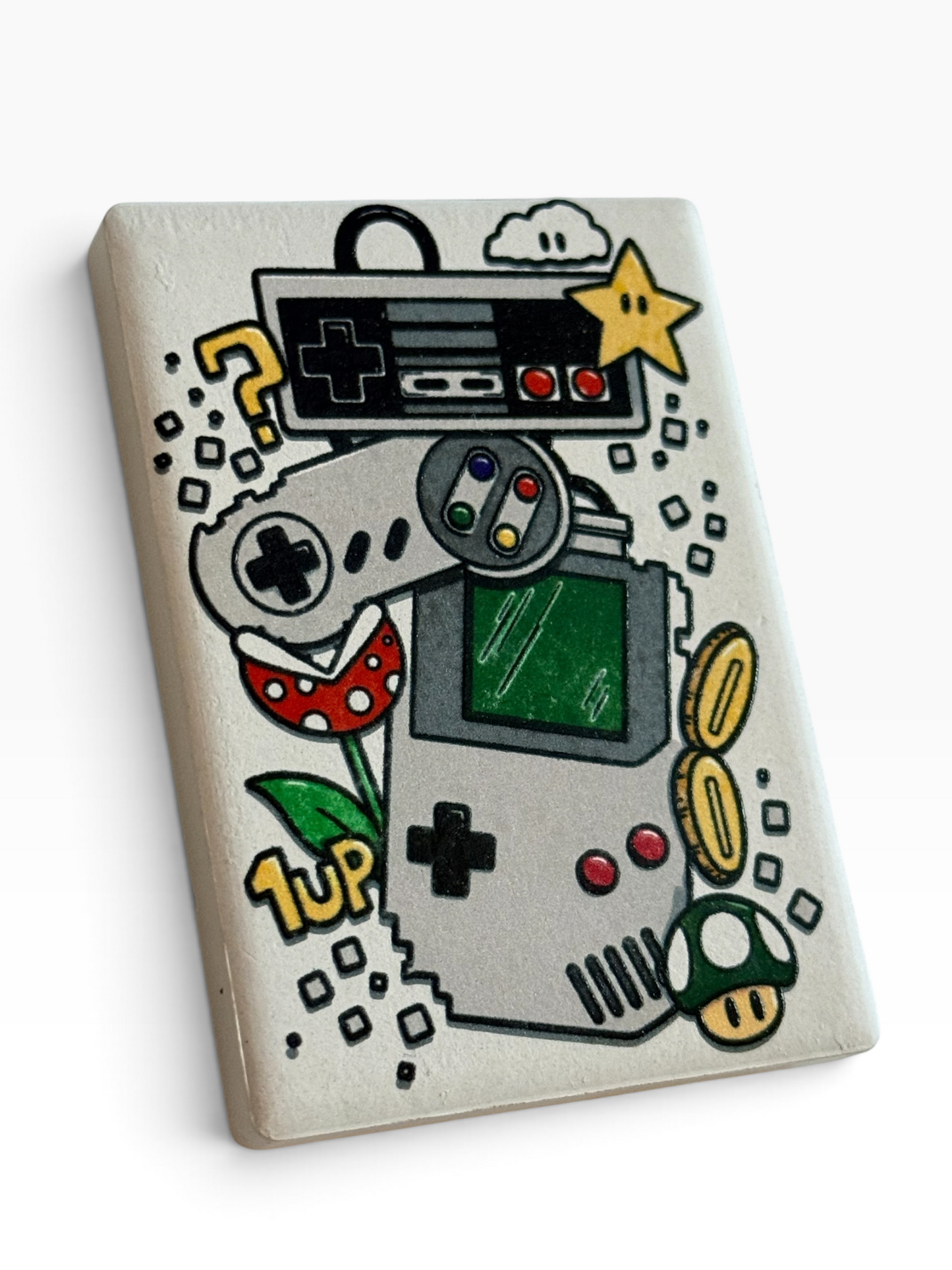A rectangular ceramic magnet featuring a colorful retro video game console illustration with a game counsel, controllers, star, coins, mushroom, and question marks on a white background.