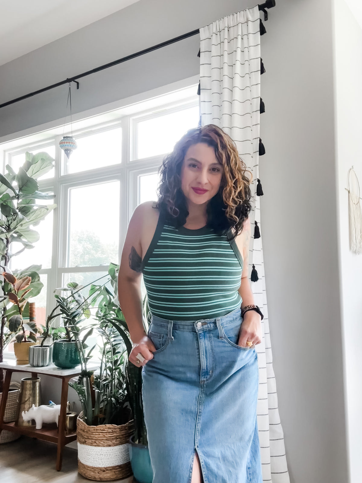 Woman in a striped top and denim skirt standing in a room with plants and a window.