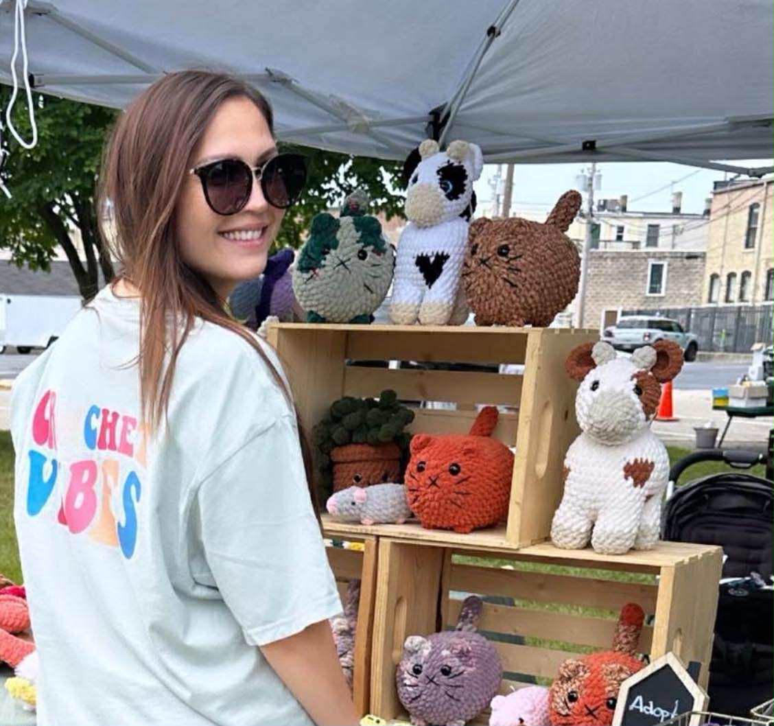 Woman standing next to a display of crocheted animals at an outdoor event.