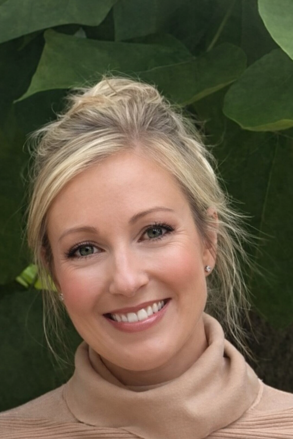 Woman with blonde hair and a beige sweater against a green leafy background