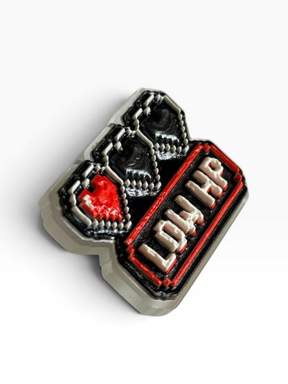 A pixel art acrylic uv printed pin featuring a red heart with a black heart outline, a red and black striped background with 3D text reading 'LOW HP', and clear base, shown on a white background with a soft shadow.