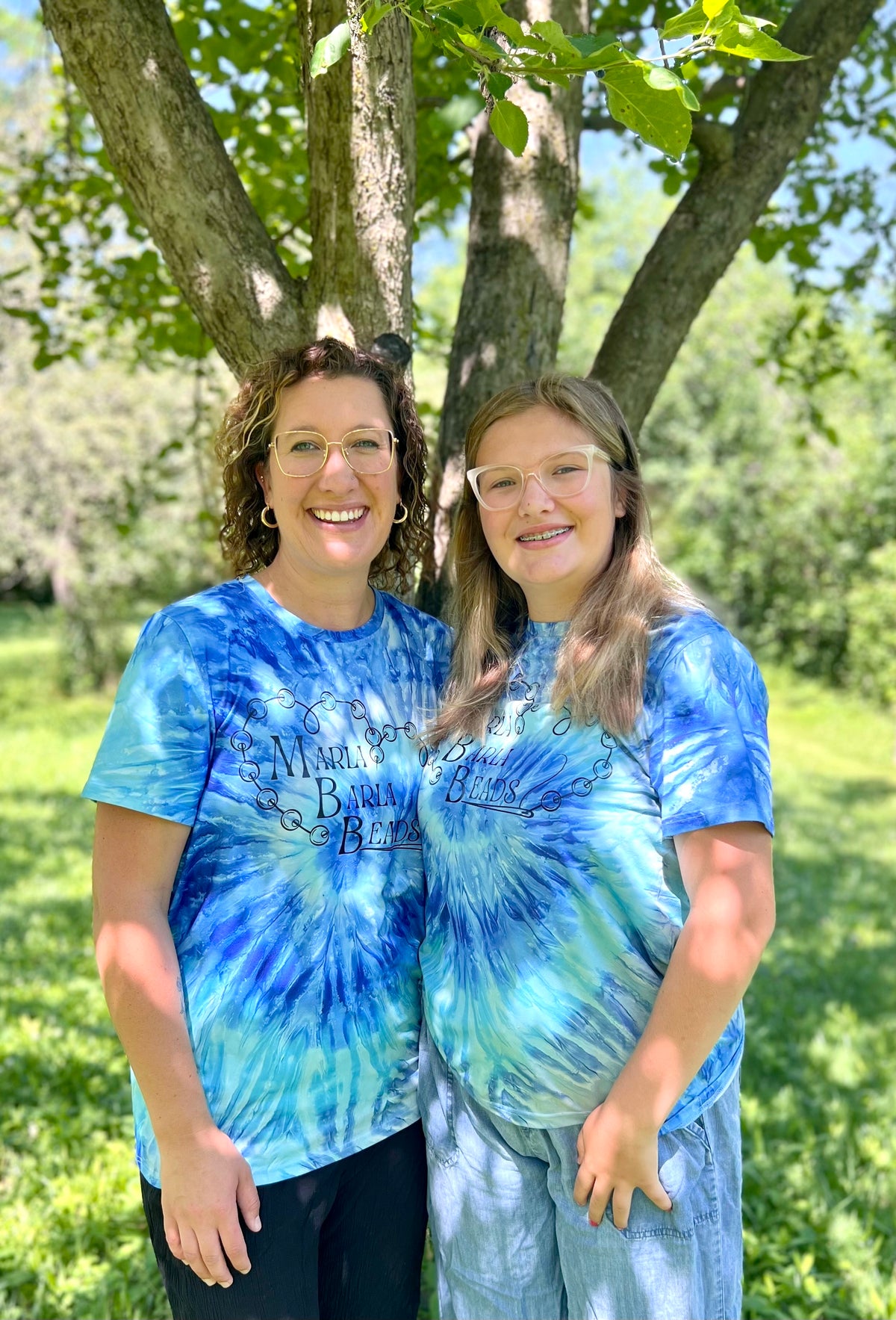 Two women wearing blue tie-dye shirts standing outdoors under a tree.