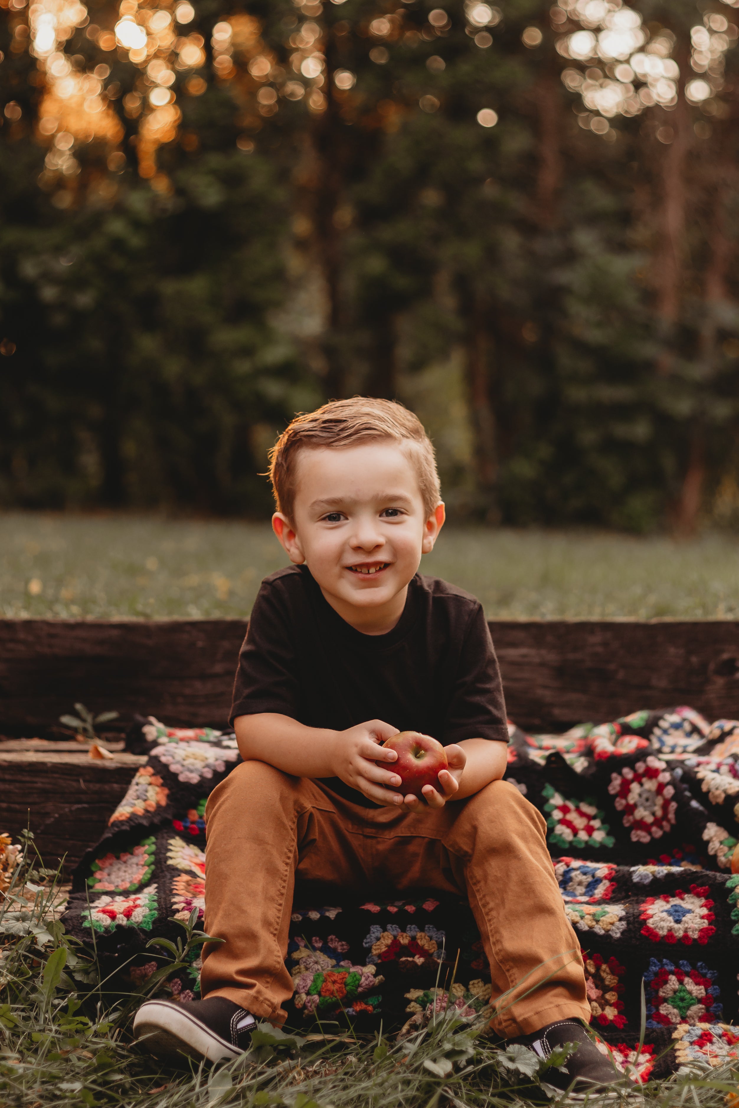 Photo of a young boy sitting on a a piece of wood and blanket holding and apple and smiling. 