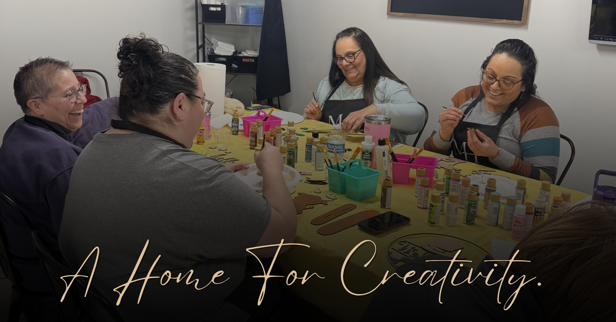 Four people seated around a table filled with craft supplies, paint bottles, and tools in an indoor room. Large script text across the bottom reads, 'A Home For Creativity.