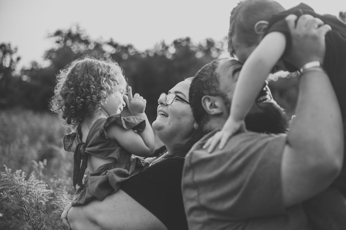 Photo of four people, two adults and two children.  A woman is holding a young girl who is wearing a dress and has curly hair. A man is holding a young boy and their foreheads are touching. 