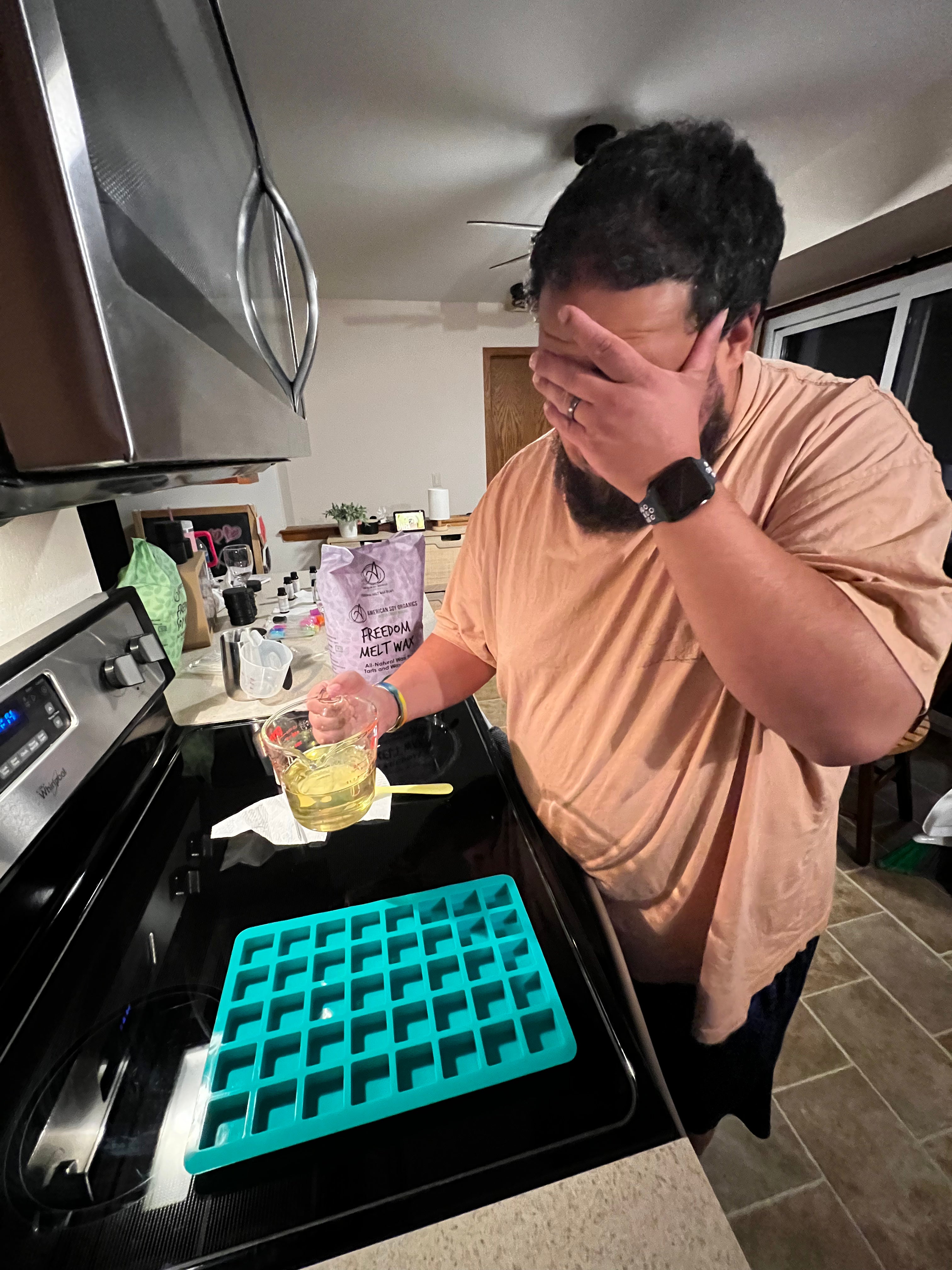 A photo of a man standing in a kitchen setting holding a measuring cup of wax and preparing to pour the contents of the glass into a silicone mold.