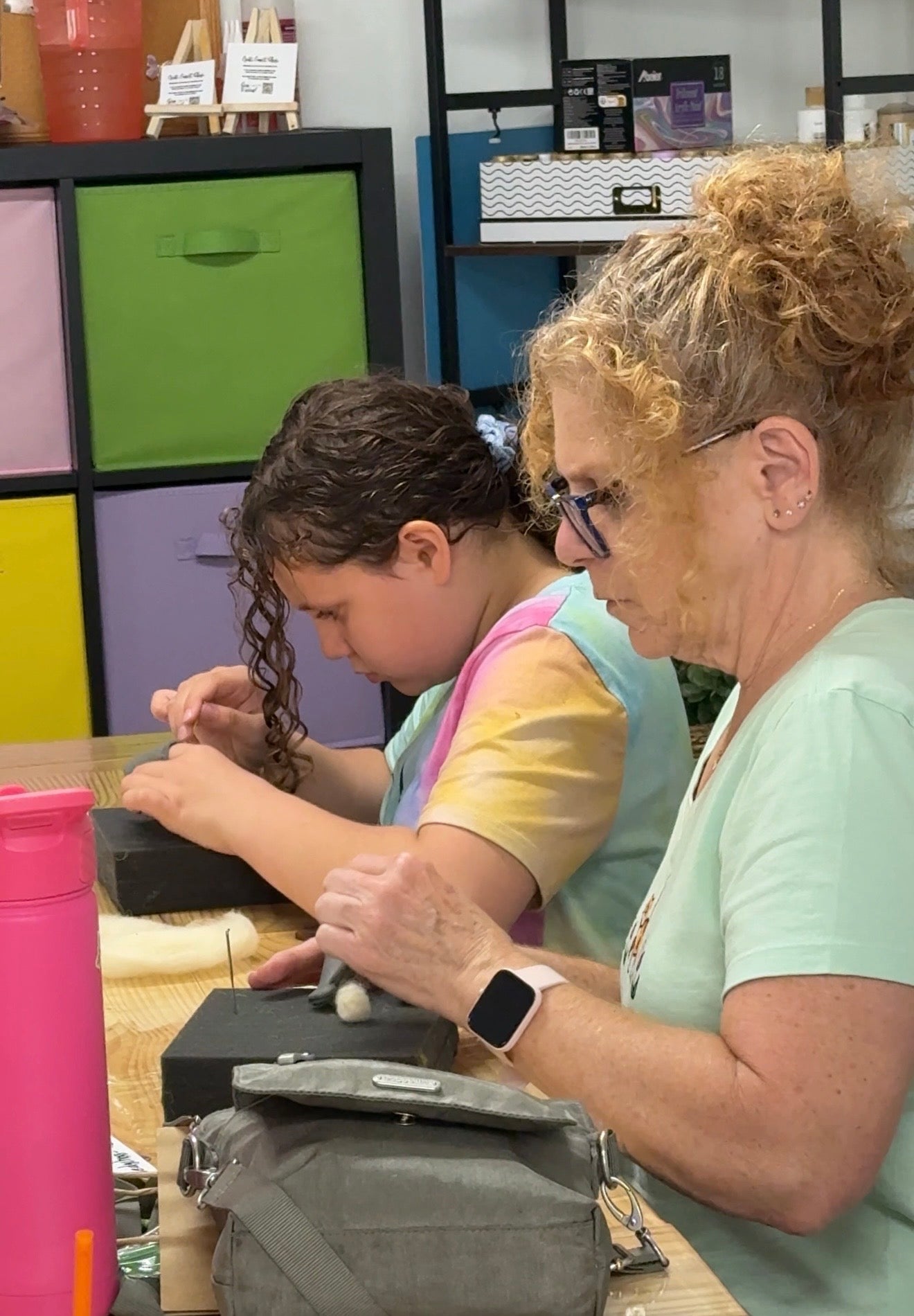 An adult woman and a young girl are seated at a table, working on small needle felting craft projects with black blocks in a colorful indoor studio or classroom setting.