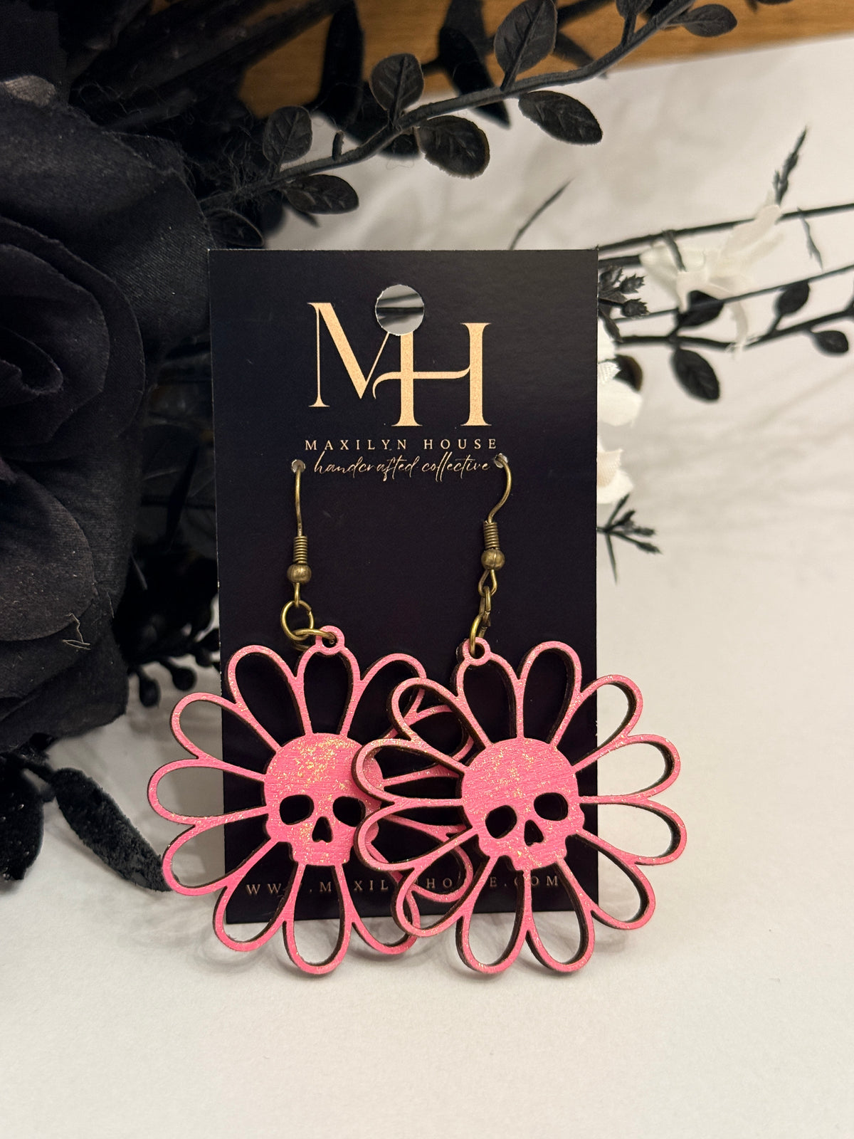 Pink laser-cut daisy earrings with skull centers, displayed on a black card labeled 'Maxlyn House' with decorative black and white flowers in the background.