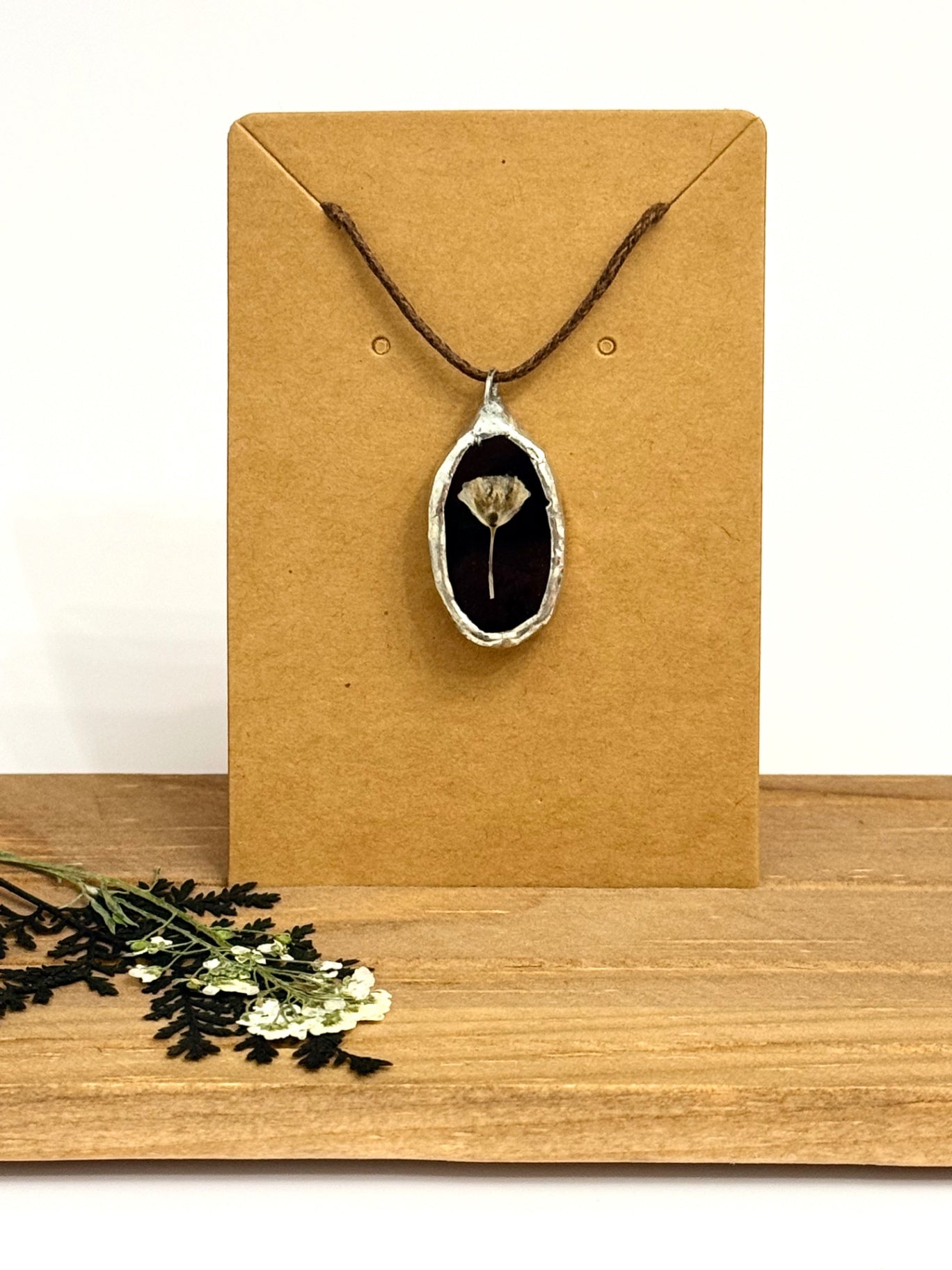 A black stained glass pendant necklace with a silver-rimmed foil setting and a dried flower inside, displayed on a brown kraft card against a white background.