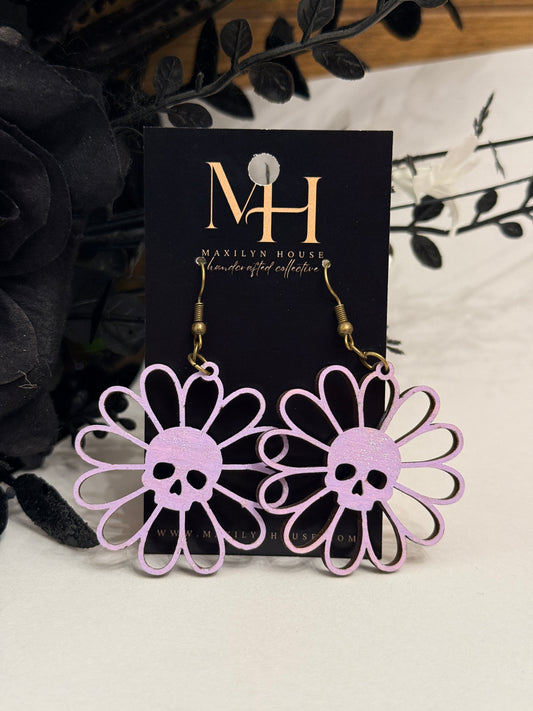 A pair of lavender daisy flower-shaped dangle earrings with skull cutouts, displayed on a black card with gold lettering that reads 'MH' and 'Maxilyn House Handcrafted Collection'. The background is styled with black artificial roses and dark leaves on a light beige surface.