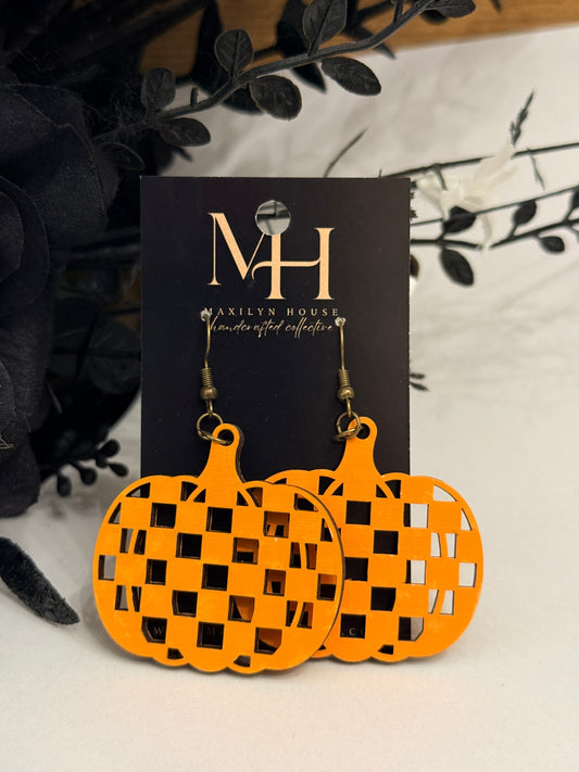 Bright orange pumpkin-shaped dangle earrings with a checkerboard cutout pattern, displayed on a black card labeled MH. The earrings are styled with dark decorative leaves on a light surface.