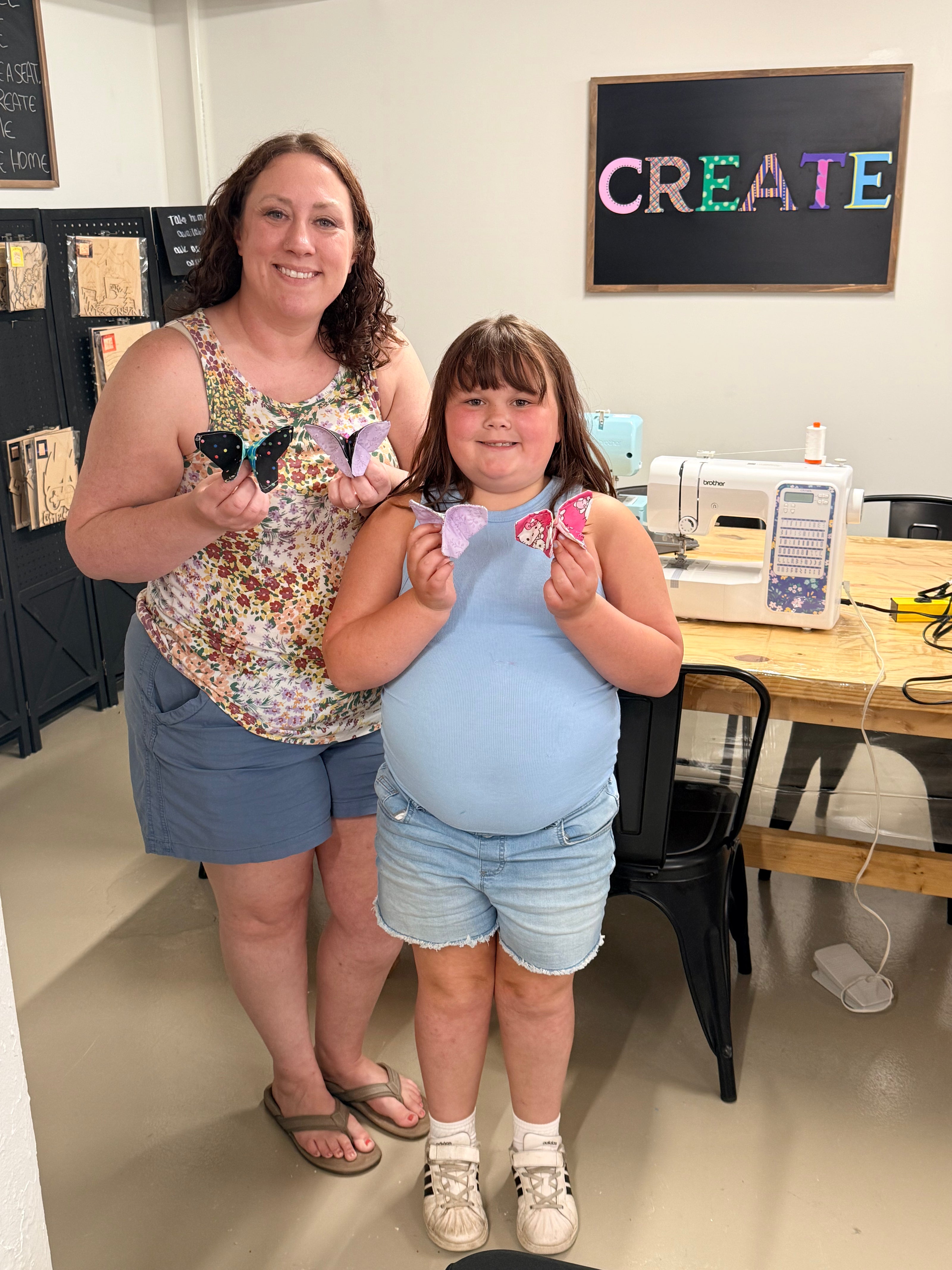 A woman and a young girl standing together in a craft class room, each holding a sewn handmade butterfly. The room has a sewing machine on a table, a chalkboard with the word 'CREATE' in colorful letters, and shelves on the left.