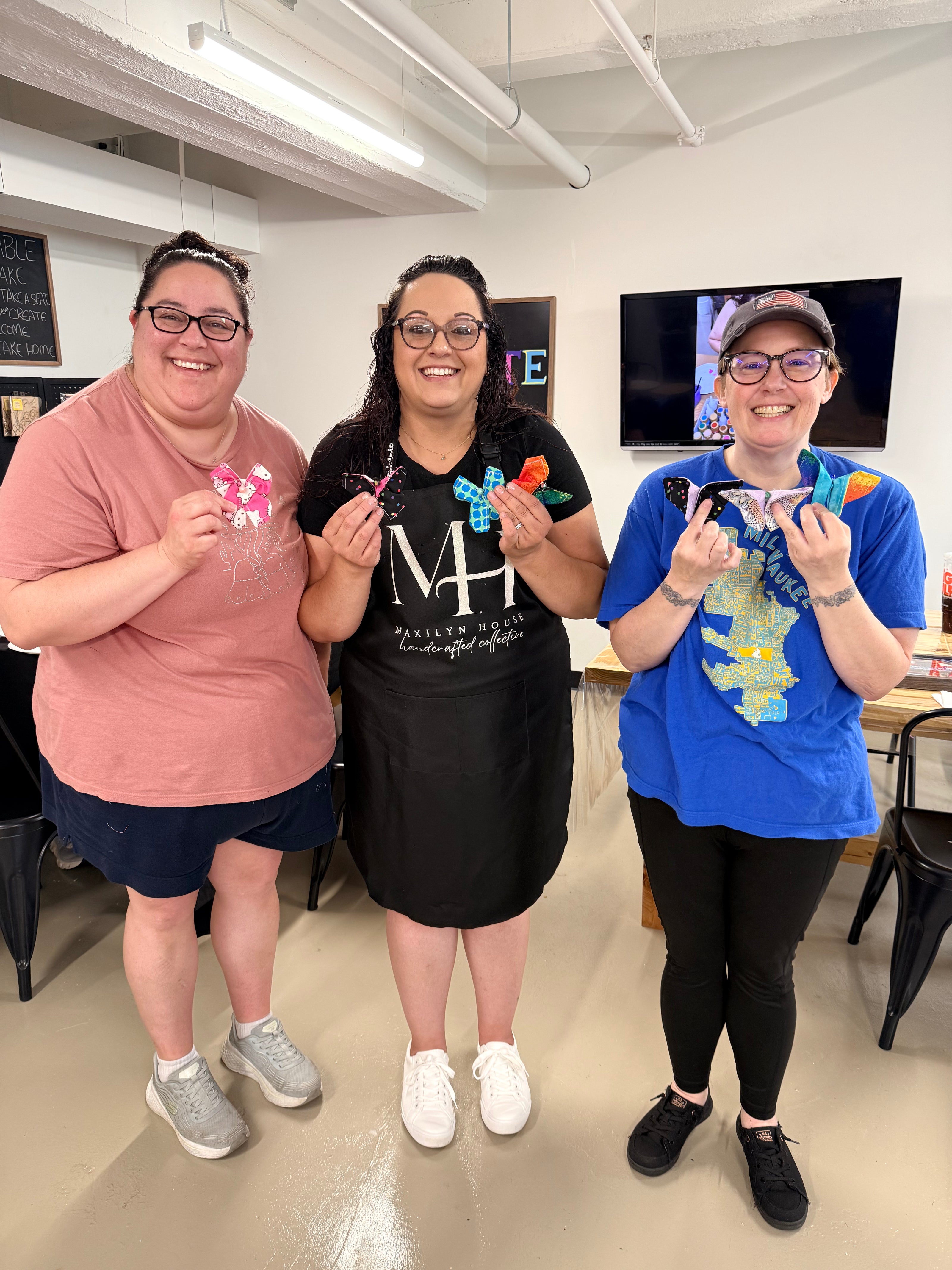 Three women standing together in a bright indoor room, each holding up a completed small sewn butterfly from their sewing class. They are wearing casual clothing, some have art smocks on. Set in a workshop classroom setting.