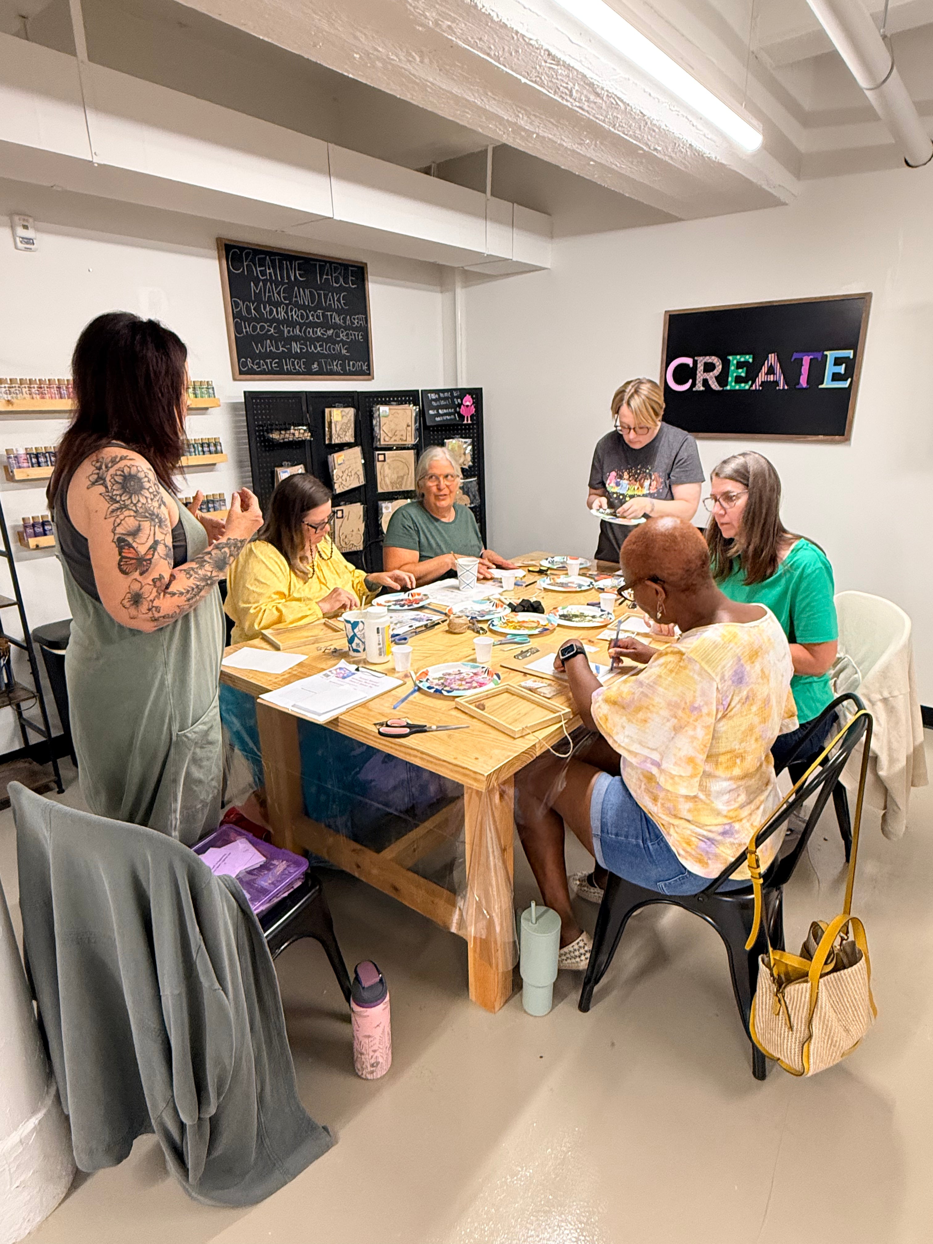A group of five women gathered around a wooden craft table in a bright studio space, engaged in a floral art frame workshop. One woman with tattoos stands on the left speaking, while the others sit or stand at the table with paint palettes, brushes, and mugs.