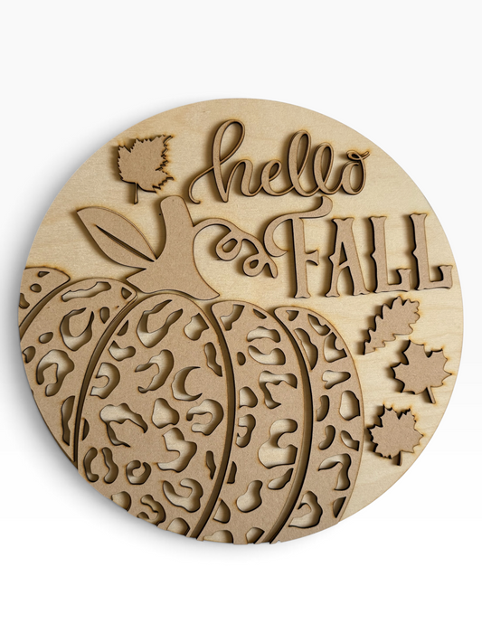 A round wood and mdf sign featuring a carved pumpkin with a leopard print pattern, the words 'hello and FALL' in stylized lettering, and several small leaf cutouts, shown on a plain white background.