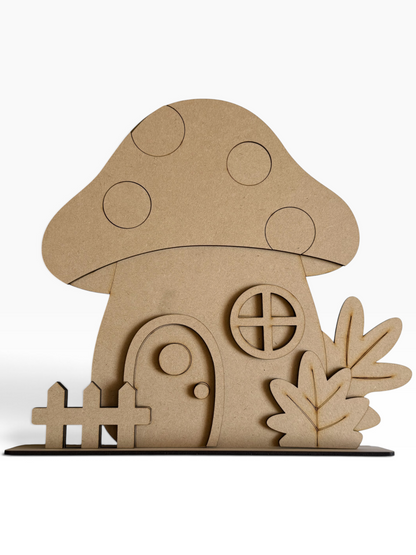 A fall themed unfinished MDF decorative cutout of a mushroom house with a small fence and leafy plants, shown on a clean white background.