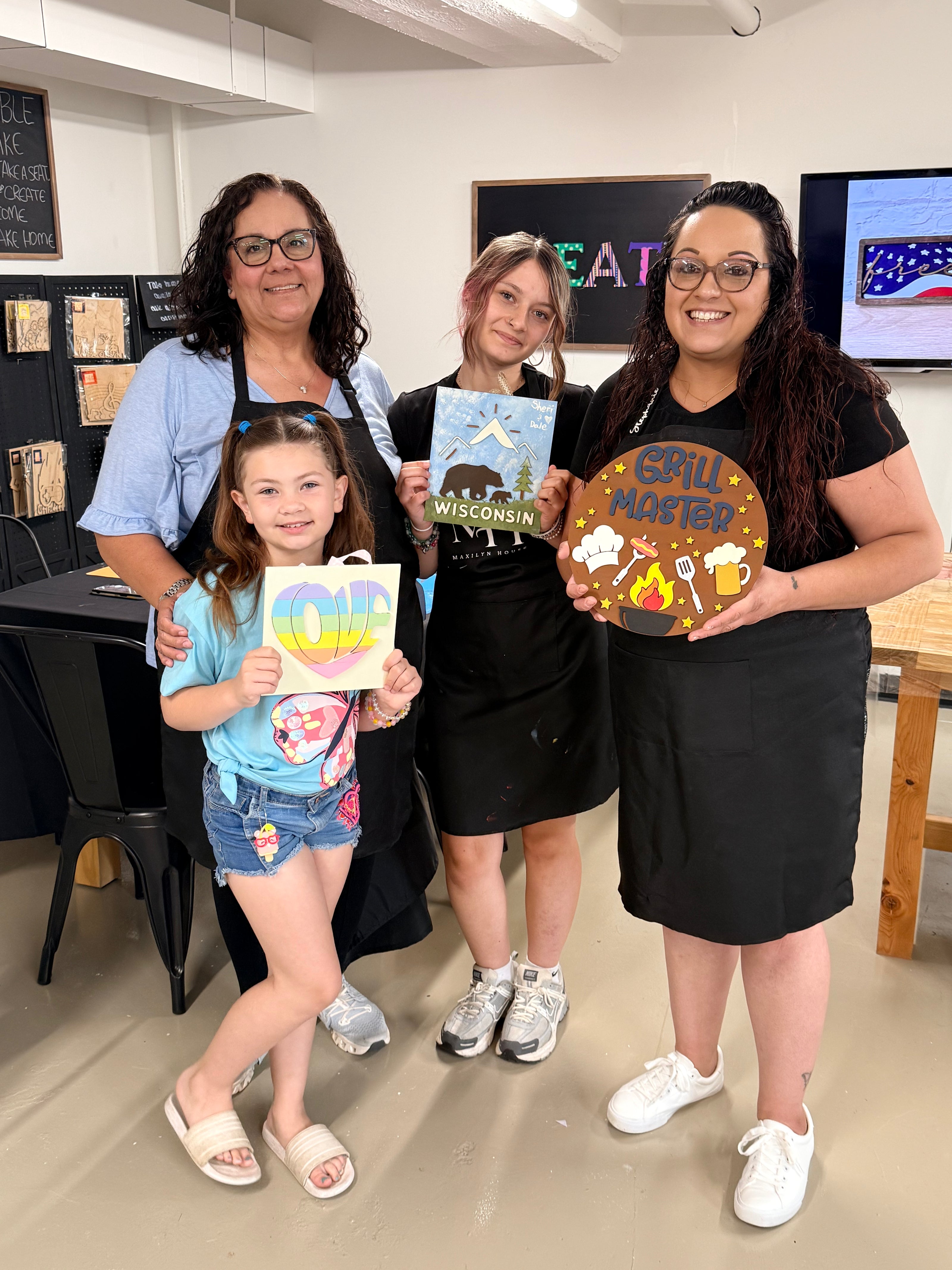 Four people standing together in a workshop setting, each holding a colorful painting project. The group includes two women and two children, and the background shows desks, blackboards, and a screen.
